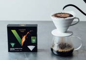 Eine schwarze Schachtel Hario V60 Meteor Kaffee-Papierfilter steht auf einer grauen Oberfläche neben einem Pour-Over-Kaffee-Set. Aus einem schwarzen Schwanenhals-Wasserkocher wird heißes Wasser auf das Kaffeepulver in einem weißen Keramik-Dripper gegossen, während der frisch gebrühte Kaffee in eine Glaskanne darunter tropft.