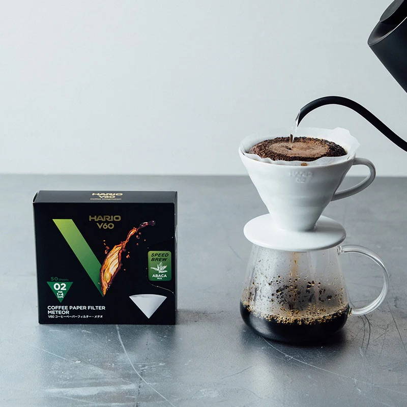 A black box of Hario V60 Meteor coffee paper filters sits on a gray surface next to a pour-over coffee setup. Hot water is being poured from a black gooseneck kettle onto coffee grounds in a white ceramic dripper, brewing coffee into a glass carafe below.