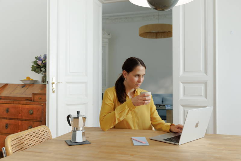 Frau sitzt mit Laptop am Tisch und hält Kaffeebecher