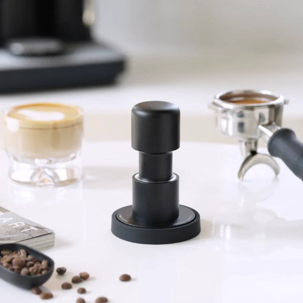 Der schwarze Timemore Espresso-Tamper steht aufrecht auf einer weißen Oberfläche. Im unscharfen Hintergrund sind ein Siebträger, eine Tasse Cappuccino und gestreute Kaffeebohnen zu sehen.