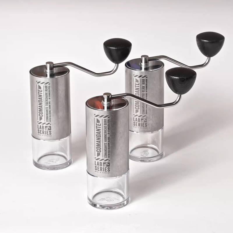 Produktfoto von drei Comandante C40 LAB Series Handkaffeemühlen auf weißem Hintergrund. Die Mühlen haben ein Gehäuse aus nacktem, industriell anmutendem Edelstahl, Kurbeln mit schwarzen Holzknäufen und klare Polymer-Auffangbehälter. Sie sind leicht versetzt aufgestellt.