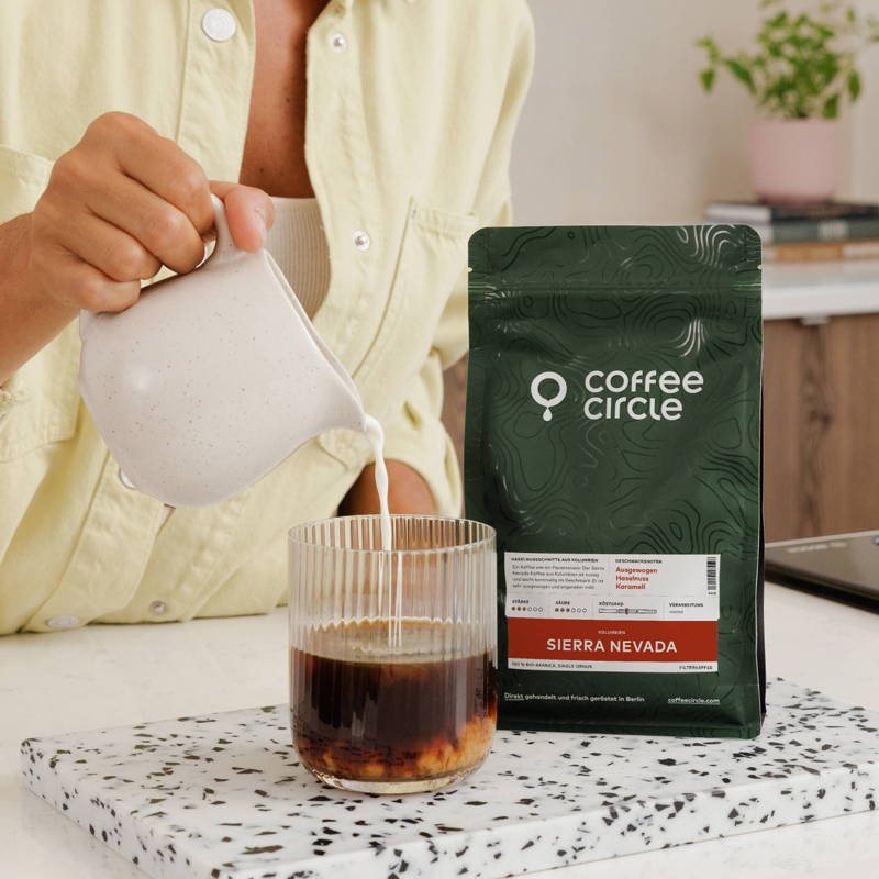 A person pouring milk from a small speckled pitcher into a ribbed glass of black coffee, showing the liquids swirling together. Next to the glass stands a dark green bag of "Coffee Circle" coffee labeled "Sierra Nevada," placed on a black and white terrazzo board.