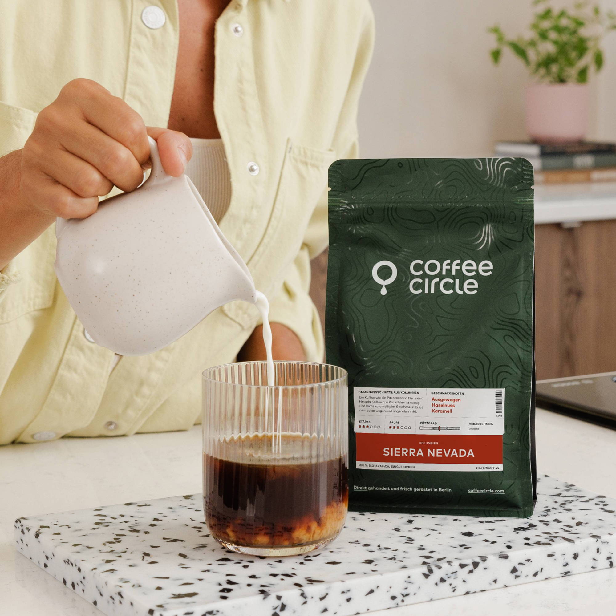 Eine Person gießt Milch aus einem kleinen, gesprenkelten Kännchen in ein geriffeltes Glas mit schwarzem Kaffee, wobei sich die Flüssigkeiten vermischen. Daneben steht eine dunkelgrüne Kaffeepackung der Marke "Coffee Circle" mit der Aufschrift "Sierra Nevada" auf einem weiß-schwarz gesprenkelten Brett.