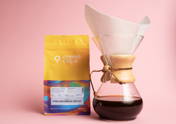 Eine Pink Bourbon Decaf Kaffeeverpackung steht neben einer Chemex mit Papierfilter vor einem rosafarbenen Hintergrund