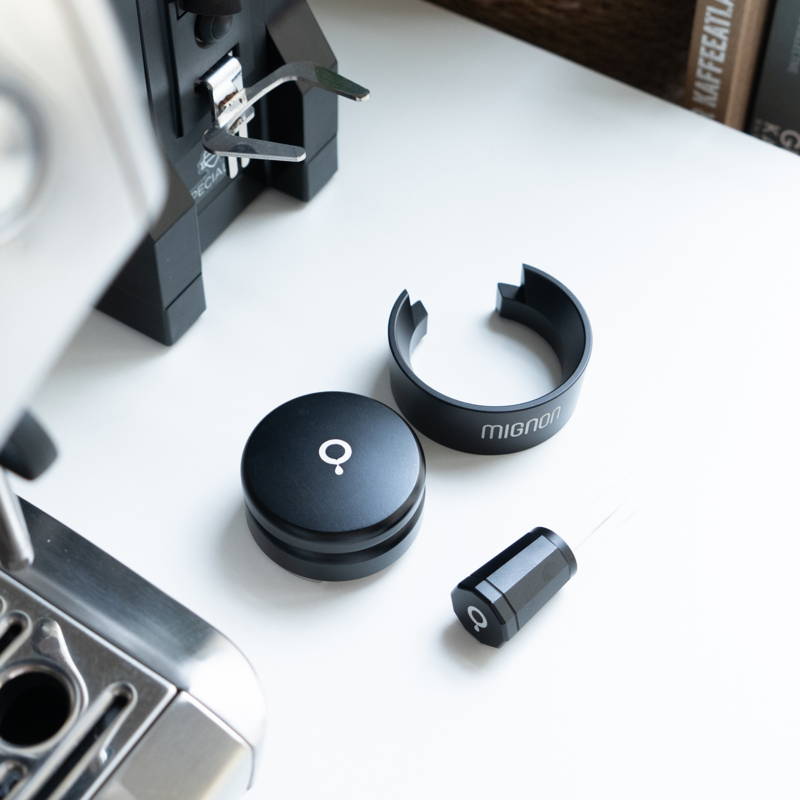 Coffee Circle Leveler, WDT-Tool und Eureka Dosing Funnel liegen auf einem weißen Tisch. Dahinter ist eine Eureka Specialità zu sehen. Ein Kaffeeatlas grenzt an den Tisch an.