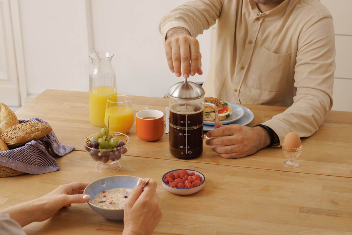 Mann brüht am Frühstückstisch Kaffee mit der French Press, Frau löffelt Müsli