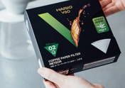 Two hands are holding a black box of Hario V60 Meteor coffee paper filters towards the camera. The box features a large green "V", the text "Speed Brew Abaca Mix", and the size "02". A blurred white ceramic coffee dripper is visible in the background.
