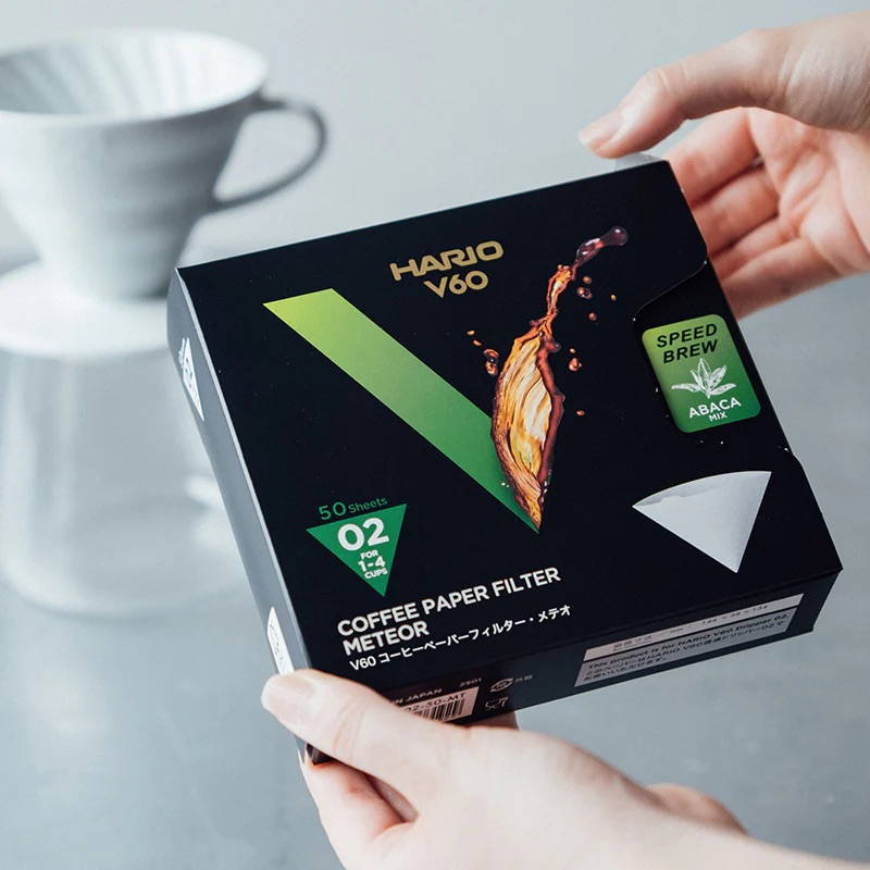 Two hands are holding a black box of Hario V60 Meteor coffee paper filters towards the camera. The box features a large green "V", the text "Speed Brew Abaca Mix", and the size "02". A blurred white ceramic coffee dripper is visible in the background.