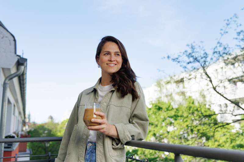 Frau steht auf Balkon und hält Iced coffee in der Hand