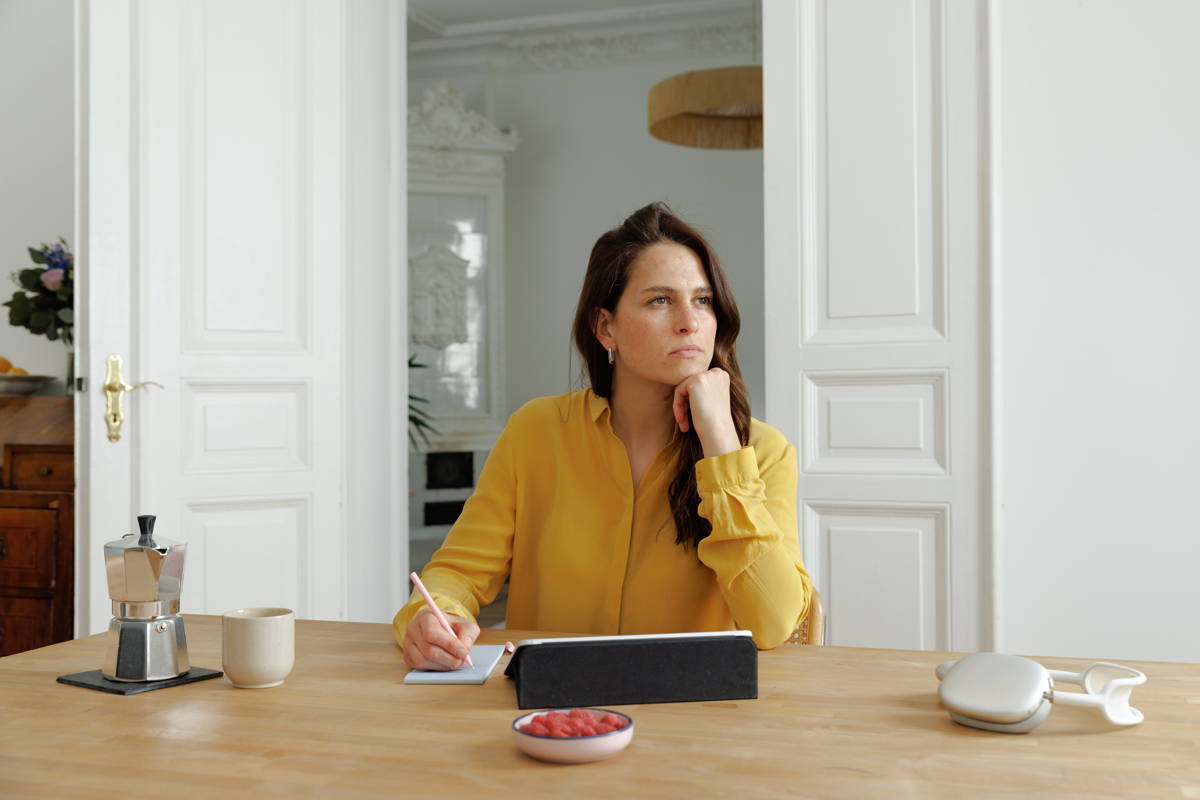 Frau sitzt mit Laptop am Tisch und schaut nachdenklich während sie etwas aufschreibt