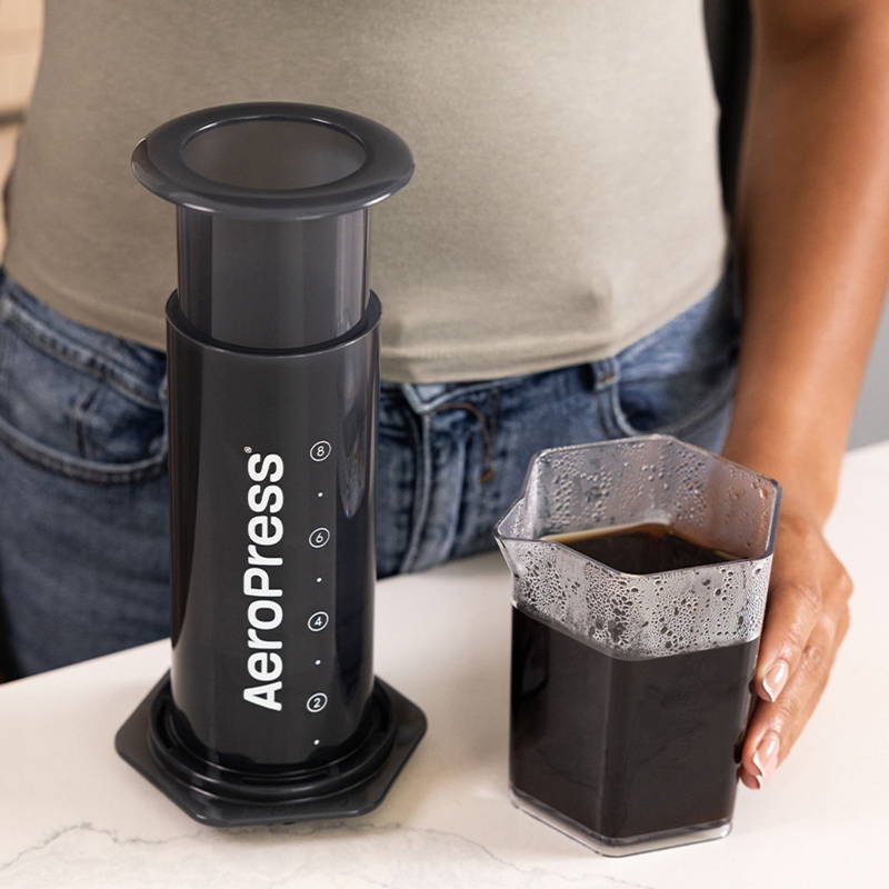 AeroPress XL steht auf einem Tisch. Daneben das mit Kaffee gefüllte AeroPress Trinkgefäß.