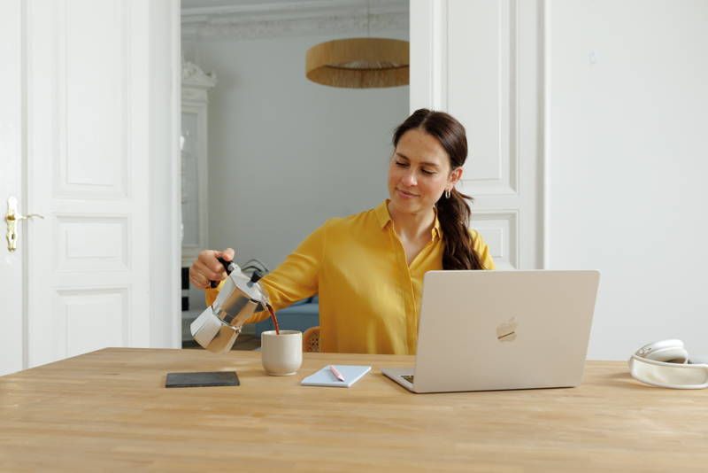 Frau sitzt mit Laptop am Tisch und gießt sich Kaffee aus Bialetti in Kaffeebecher