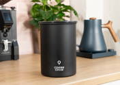 Schwarze Kaffeedose mit weiße Logo von Coffee Circle auf einer hölzernen Küchenarbeitsplatte, im Hintergrund eine Kaffeemühle, eine Zimmerpflanze und ein elektrischer Wasserkocher.