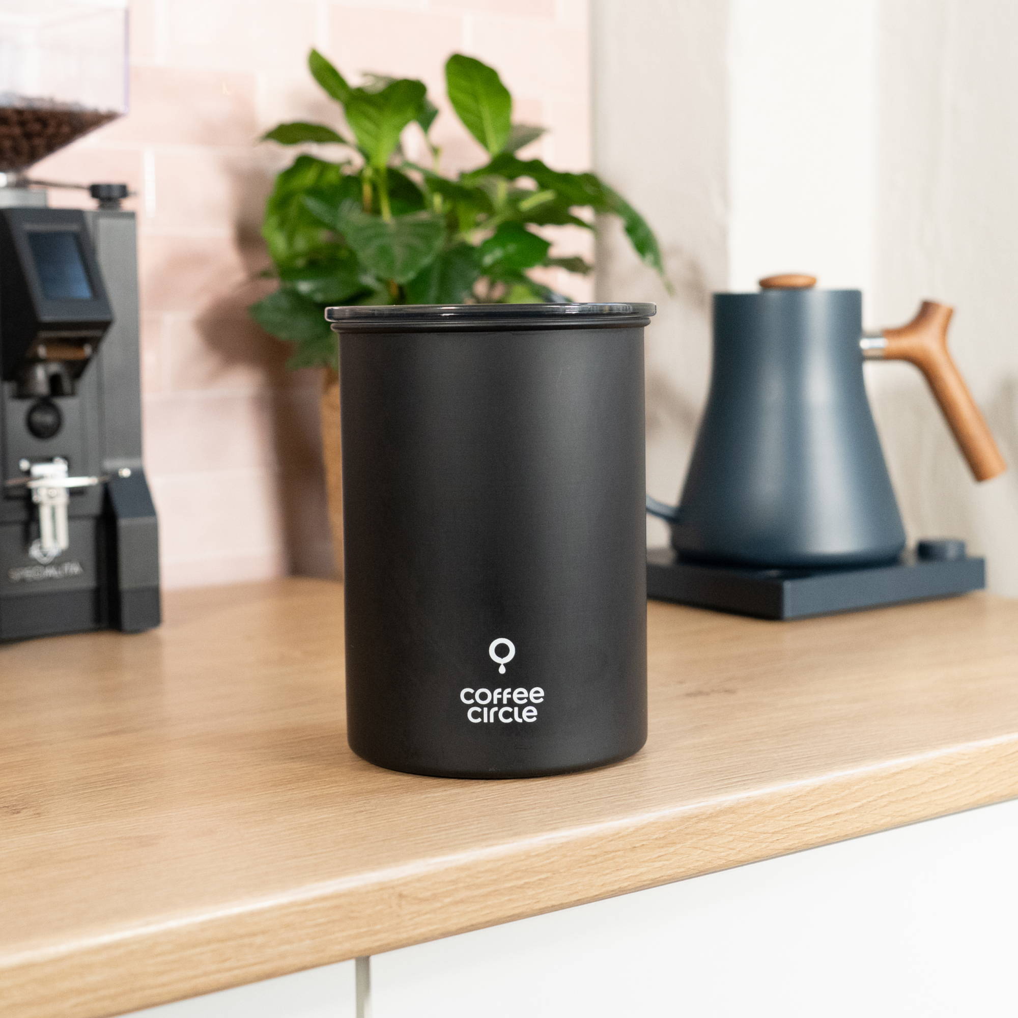 Cylindrical matte black Coffee Circle coffee canister standing on a light wooden countertop. In the blurred background, a black coffee grinder, a green potted plant, and a dark blue gooseneck kettle with a wooden handle are visible against pink tiled walls.
