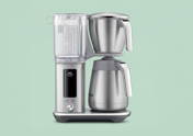Eine Sage Precision Brewer Kaffeemaschine aus gebürstetem Edelstahl auf einem hellgrünen Hintergrund. Sie verfügt über einen transparenten Wassertank mit Mengenskala (links), ein oberes Brühmodul mit Filterkorb und Griff, und darunter eine Edelstahl-Thermoskanne mit Ausgießer. Ein zentrales Display zeigt die Optionen 'BREW COLD BREW CUSTOM' mit einem leuchtenden runden Knopf darunter.