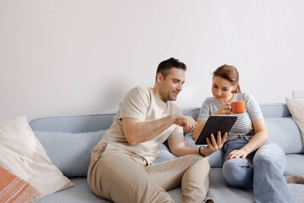 Mann und Frau sitzen auf der Couch, der Mann zeigt etwas auf dem Tablet, die Frau hält eine Kaffeetasse in der Hand