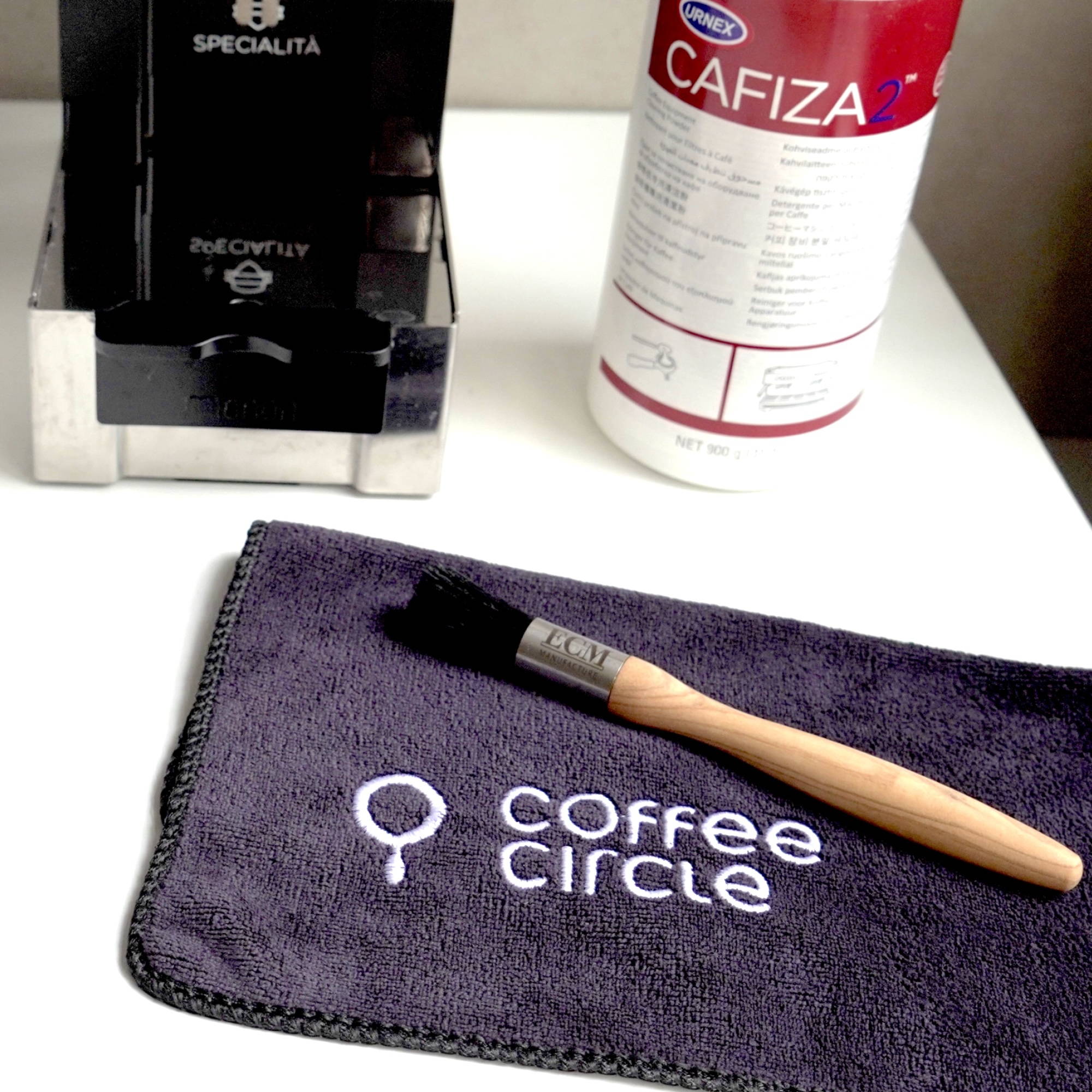Reinigungsset für Espressomaschinen auf einem weißen Tisch neben einer Kaffeemühle mit Kaffeebohnen. Auf dem Tisch liegen ein Coffee Circle Mikrofasertuch mit Holzpinsel sowie eine Dose Cafiza-Reiniger. Im Hintergrund stehen eine grüne Zimmerpflanze und zwei Kaffeebücher, darunter „Guter Kaffee“ von James Hoffmann.