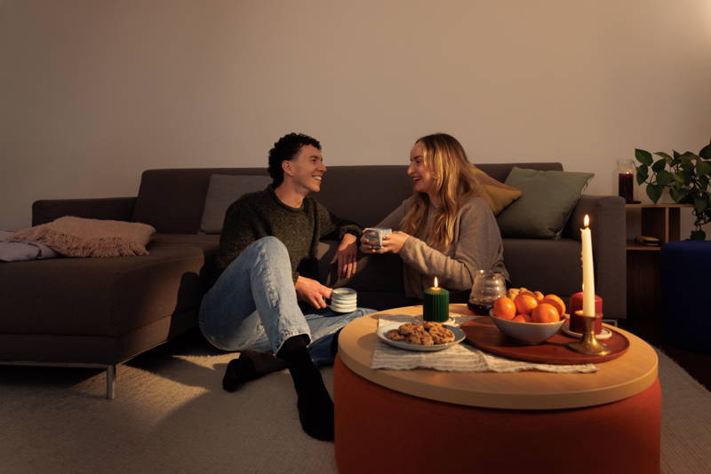 Mann und Frau sitzen kaffeetrinkend vor der Couch auf dem Boden, auf dem Tisch stehen Kerzen, Mandarinen und Kekse