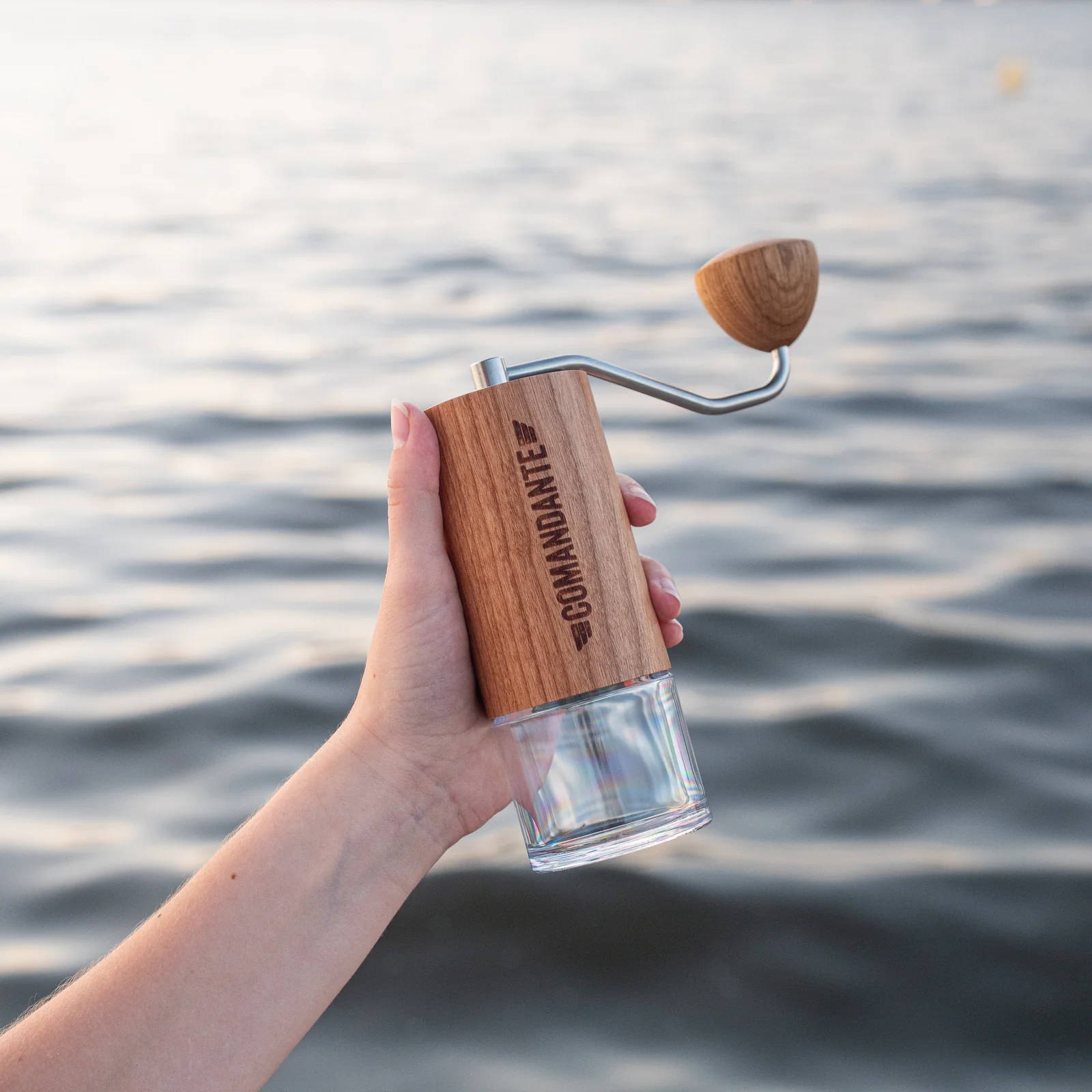 Eine Hand hält eine Comandante-Handkaffeemühle mit hellem Holzfurnier und durchsichtigem Auffangbehälter. Im unscharfen Hintergrund ist eine leicht wellige Wasseroberfläche zu sehen.