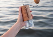 Eine Hand hält eine Comandante-Handkaffeemühle mit hellem Holzfurnier und durchsichtigem Auffangbehälter. Im unscharfen Hintergrund ist eine leicht wellige Wasseroberfläche zu sehen.