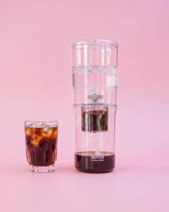 Ein Glas gefüllt mit Cold Brew Kaffee und Eiswürfeln steht links neben einem transparenten Dripster Cold Brew Zubereiter. Beide Objekte befinden sich vor einem durchgehenden, einfarbigen hellrosa Hintergrund.