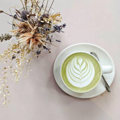 Top-down view of a white cup of matcha latte with delicate foam latte art on a saucer, next to a small glass vase with dried flowers and lavender on a pastel pink background.