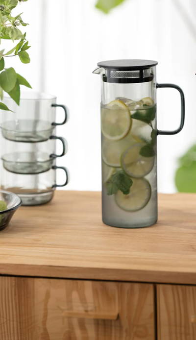 A sunlit tabletop arrangement featuring two modern glass pitchers: one amber-colored filled with tea, and one clear filled with water, limes, and mint. A stack of three smoke-grey glass cups sits next to them. In the foreground, there is a small white dish with green and dark grapes, and a single lime. The background is a bright room with white sheer curtains.