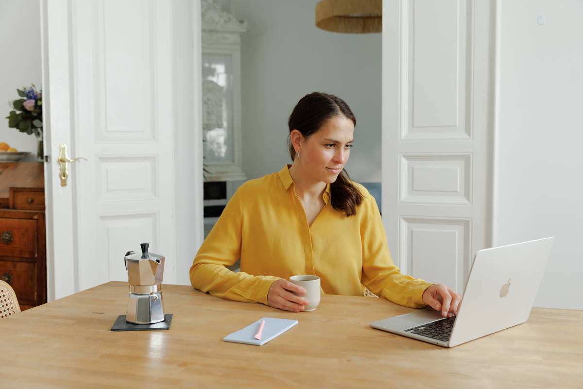 Frau sitzt mit Laptop am Tisch und hält Kaffeebecher.