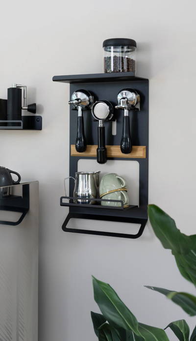 Ein moderner, schwarzer Metall-Wand-Organizer für Kaffeezubehör mit mehreren Ebenen. Er verfügt über einen oberen Boden mit einem klaren Vorratsglas für Kaffeebohnen, Halterungen für drei hängende Siebträger, eine Eichenholzleiste, ein unteres Ablagefach mit einem Edelstahl-Milchkännchen und zwei gestapelten hellgrünen Tassen sowie eine Handtuchstange ganz unten. Der Organizer befindet sich an einer hellgrauen Wand in einer sauberen Küchenumgebung.