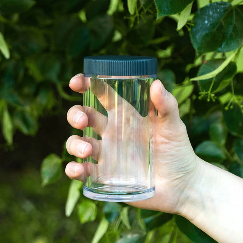 Comandante Polymer large bean container is held in the hand. Behind it, you can see a background of leaves.