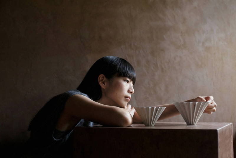 A woman gazing contemplatively at two white ceramic origami-style pour-over coffee drippers on a wooden table against a textured wall with moody lighting.