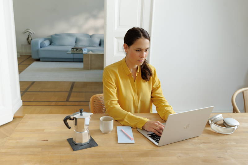 Frau sitzt mit Laptop am Tisch und hält Kaffeebecher.