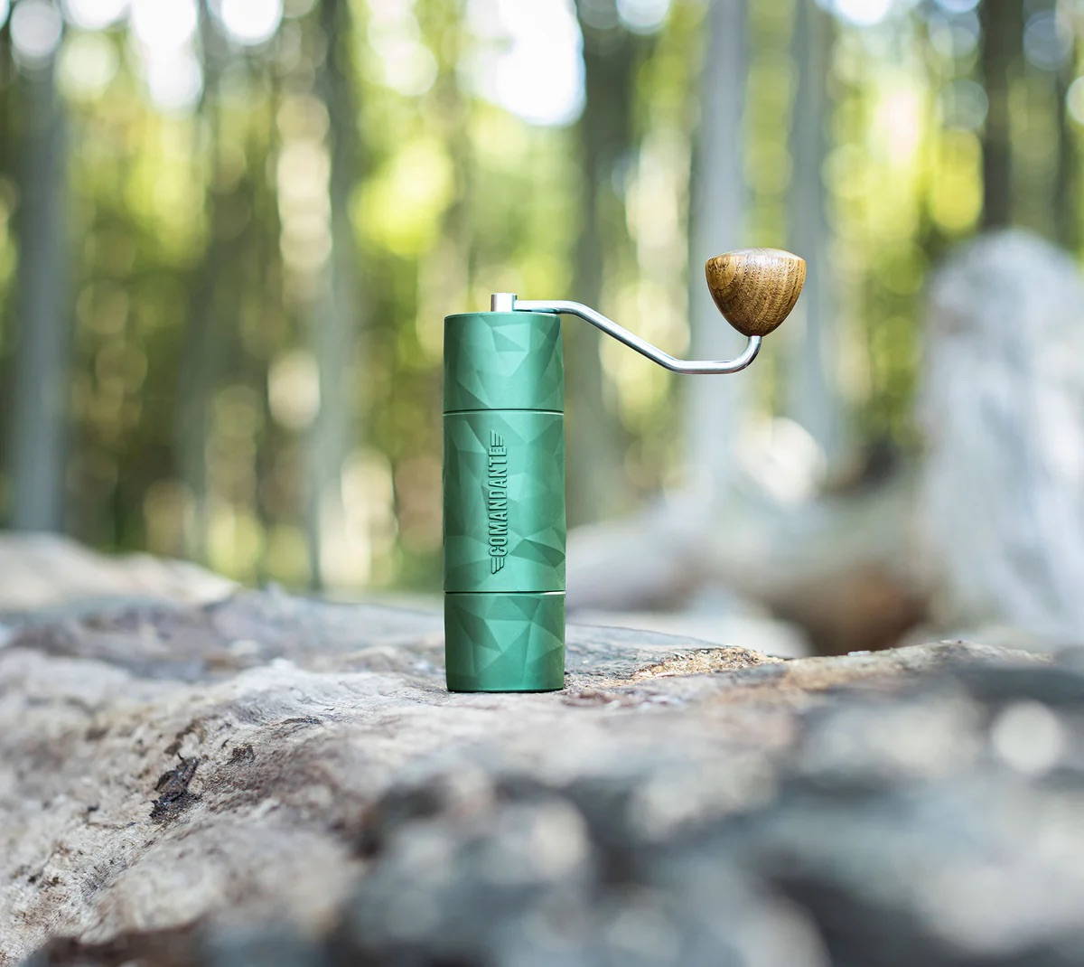 Comandante X25 Trailmaster Forest coffee grinder standing upright on a large log in a sunlit forest. The full view of the grinder with its attached metal crank arm and wooden handle shows the softly focused woodland background.
