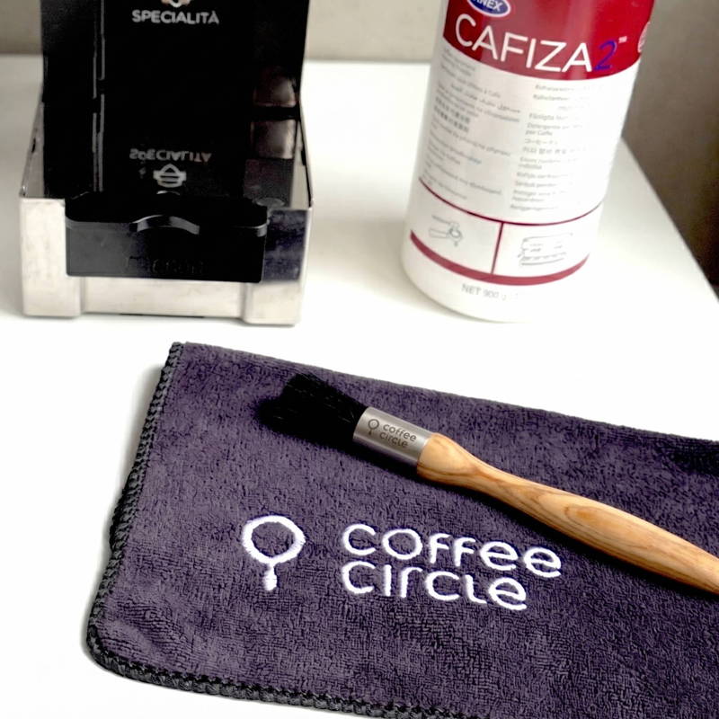 A barista cleaning set on a white table. In the foreground is a dark gray microfiber cloth with a white embroidered "coffee circle" logo, with a wooden-handled cleaning brush resting on it. In the slightly blurred background, there is a stainless steel knock box on the left and a white container of Cafiza espresso machine cleaning powder on the right.