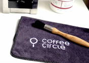 A barista cleaning set on a white table. In the foreground is a dark gray microfiber cloth with a white embroidered "coffee circle" logo, with a wooden-handled cleaning brush resting on it. In the slightly blurred background, there is a stainless steel knock box on the left and a white container of Cafiza espresso machine cleaning powder on the right.