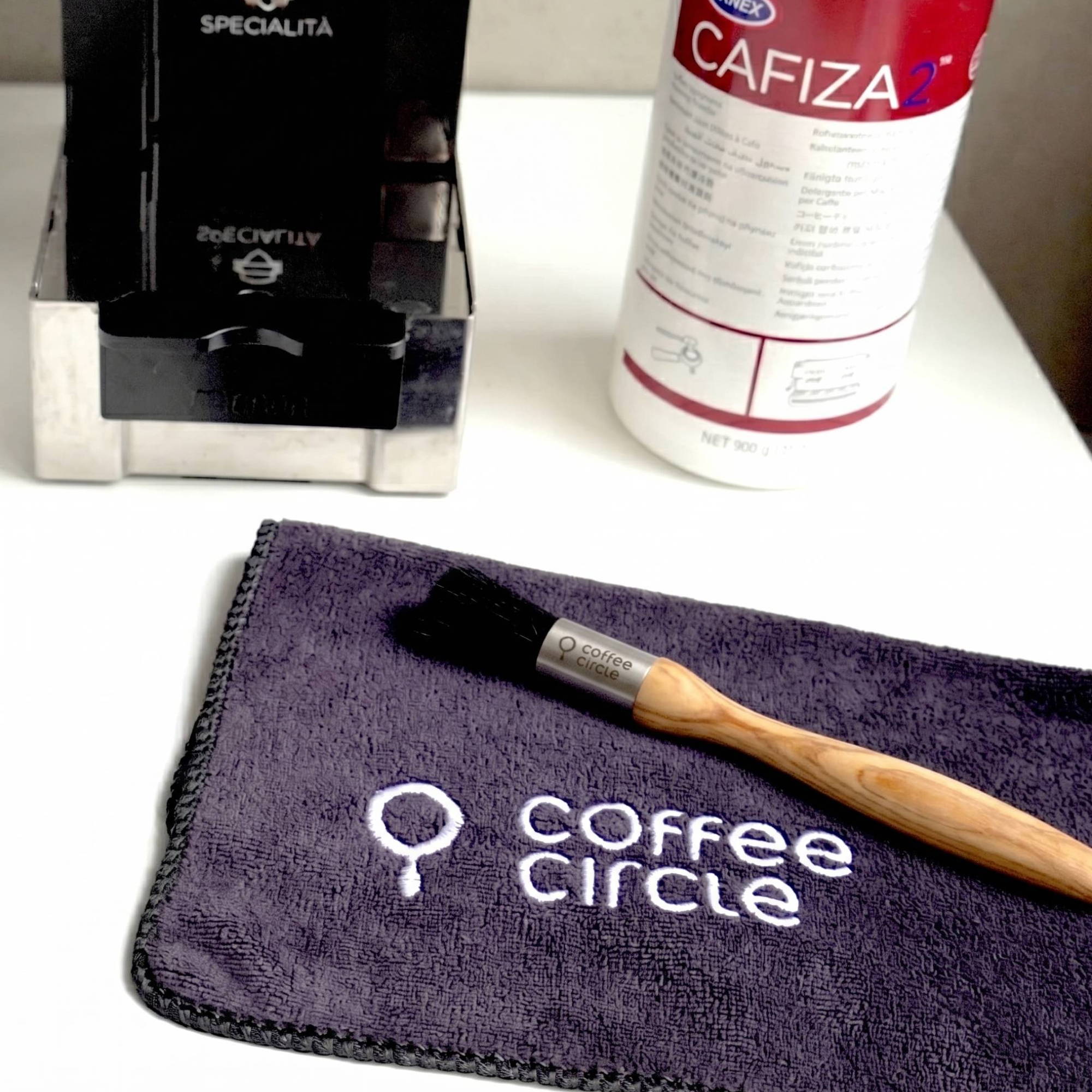 A barista cleaning set on a white table. In the foreground is a dark gray microfiber cloth with a white embroidered "coffee circle" logo, with a wooden-handled cleaning brush resting on it. In the slightly blurred background, there is a stainless steel knock box on the left and a white container of Cafiza espresso machine cleaning powder on the right.