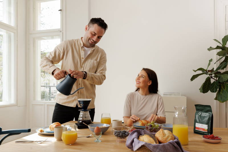 Man prepares coffee with the Hario V60 and the Fellow Stagg Pour-Over kettle, woman sits at the breakfast table and eats a croissant