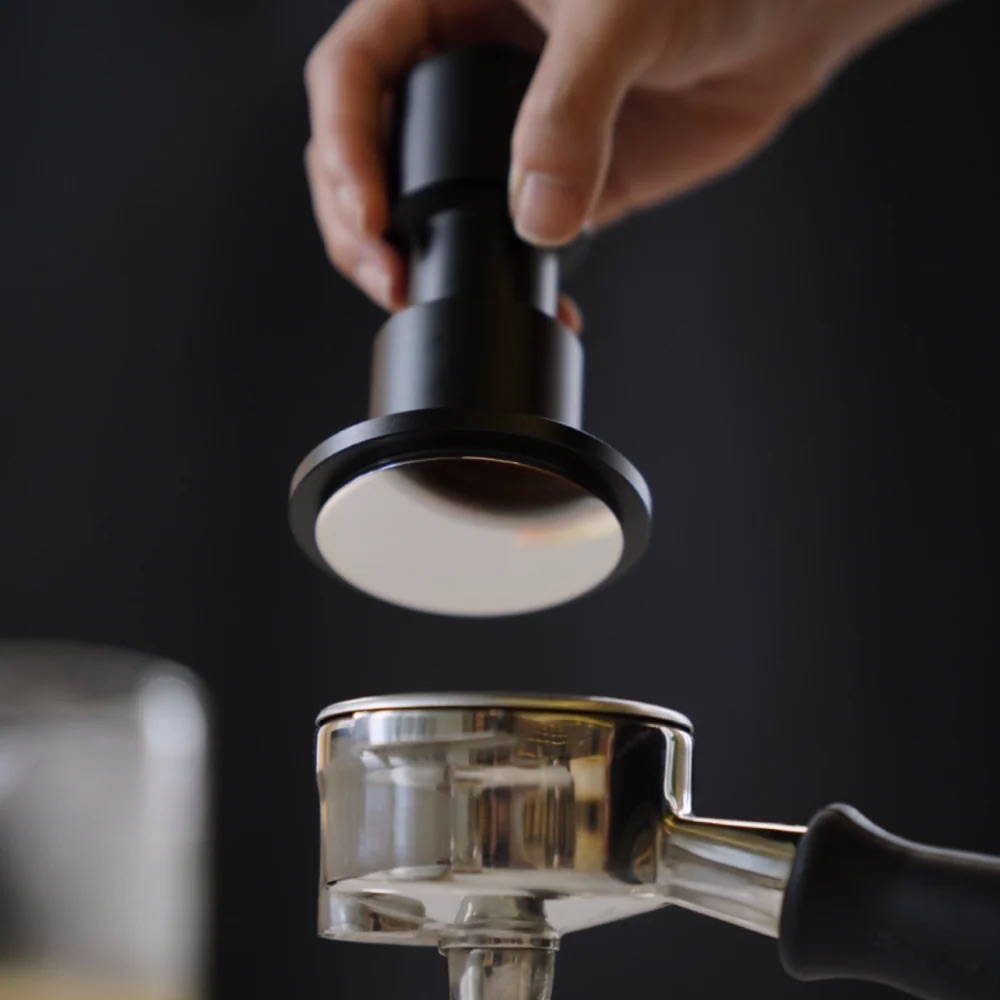 Eine Hand hält einen schwarzen Timemore Espresso-Tamper schwebend über dem Siebträger einer Kaffeemaschine vor dunklem Hintergrund.