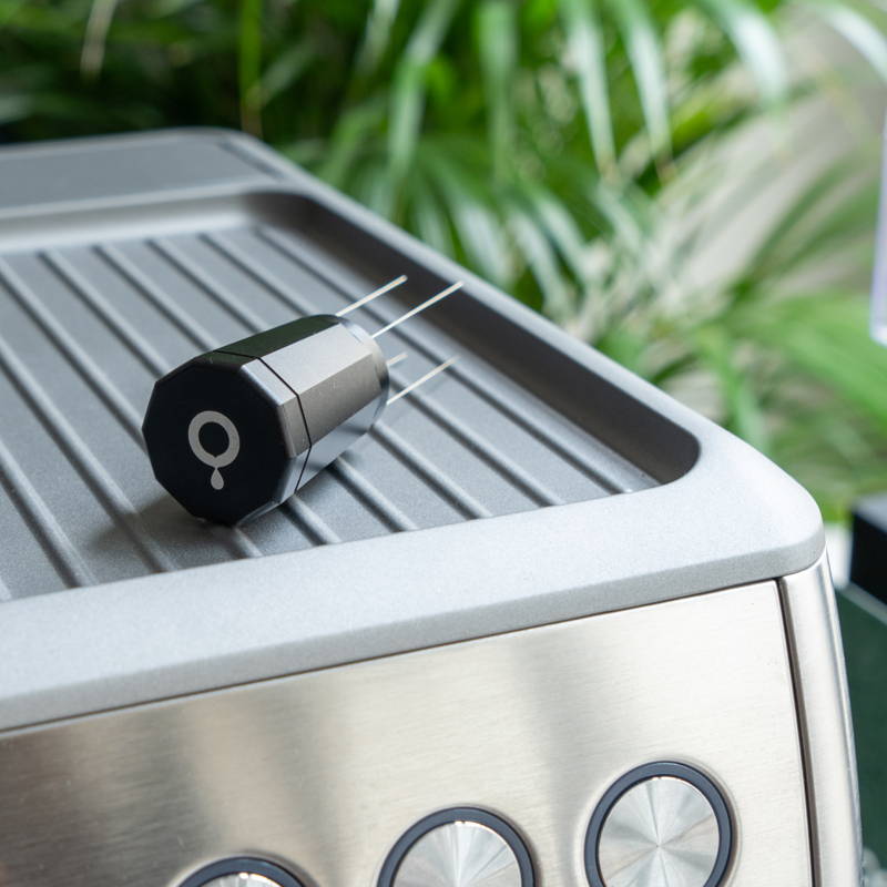 The Coffee Circle WDT tool rests on a portafilter machine. Behind it, the leaves of a plant can be seen.