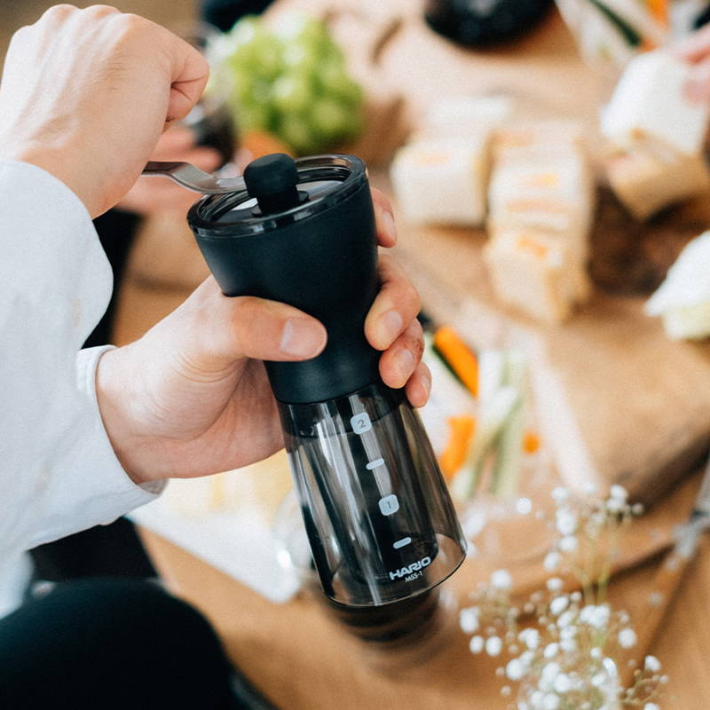 Eine Hand mahlt Bohnen in der Hario Mini Mill. Dahinter ist ein gedeckter Frühstückstisch zu sehen.