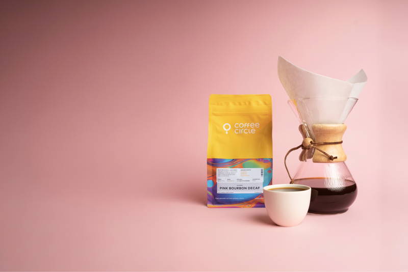 Eine Pink Bourbon Decaf Kaffeeverpackung steht neben einer Chemex mit Papierfilter und einer Tasse schwarzem Kaffee vor einem rosafarbenen Hintergrund