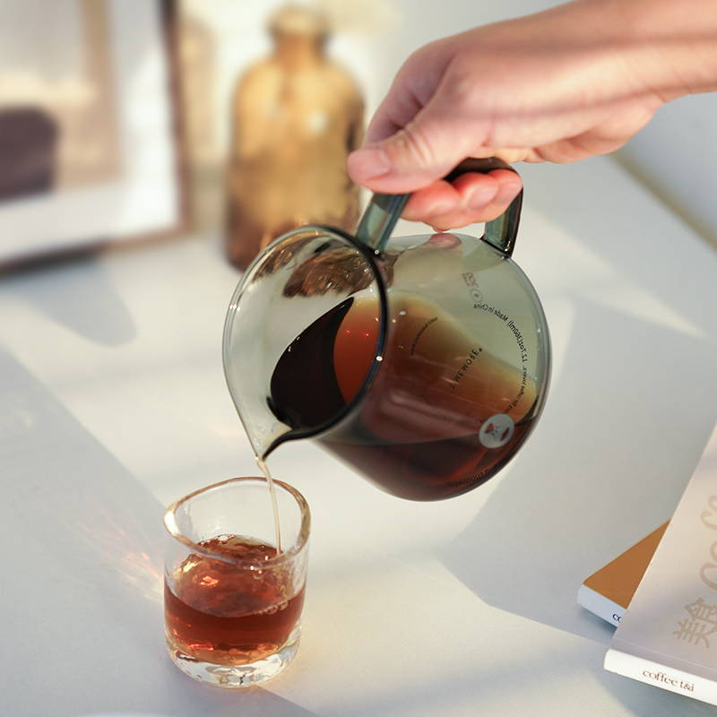 Eine Hand gießt braunen Kaffee aus einer klaren Glaskanne in ein kleines Glas auf einem weißen Tisch. Die Kanne und das Glas sind von Sonnenlicht beleuchtet und haben klare Reflexionen. Im Hintergrund befinden sich ein unscharfes Buch und eine Vase.