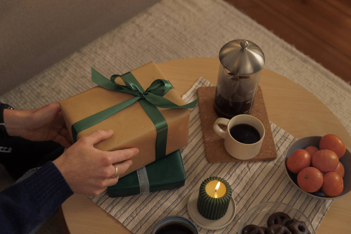 Ein Paar Hände hält zwei gestapelte, verpackte Geschenke – eines in Braun mit einer grünen Schleife und eines in Dunkelgrün mit einer weißen Schleife – neben einer Kaffeemaschine, einer brennenden Kerze und Obst auf einem Holztisch.