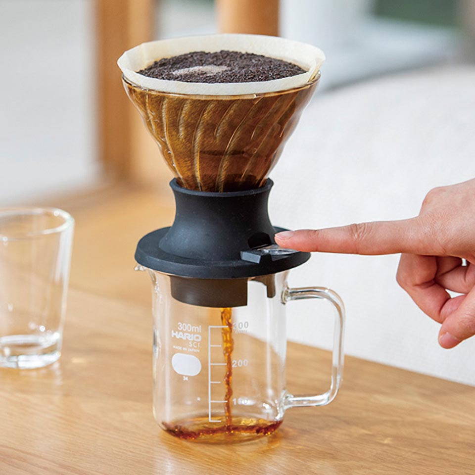 A hand pressing the black switch of an immersion coffee dripper to release the brewed coffee into the glass carafe below.