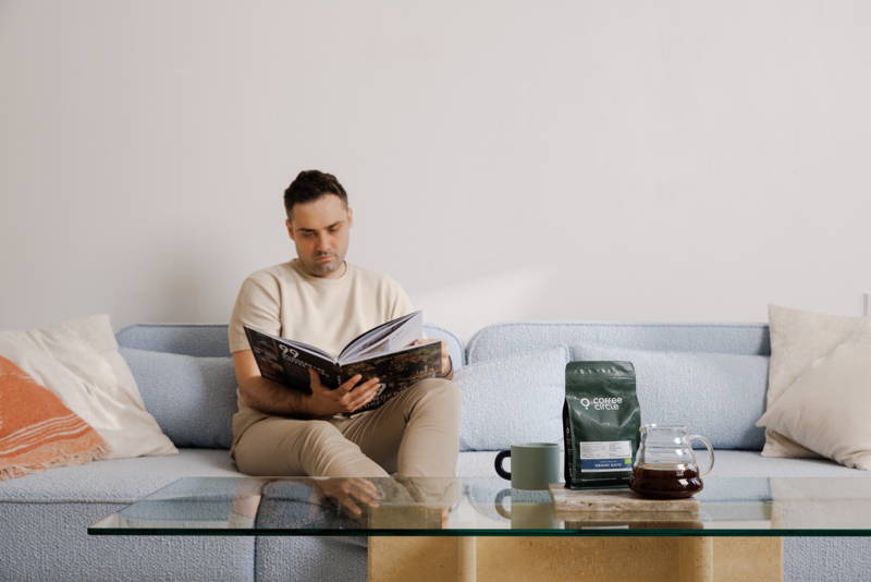 Mann liest ein Buch auf der Couch, auf dem Tisch steht eine Tasse, ein coffee bag und ein Hario V60 Range Server