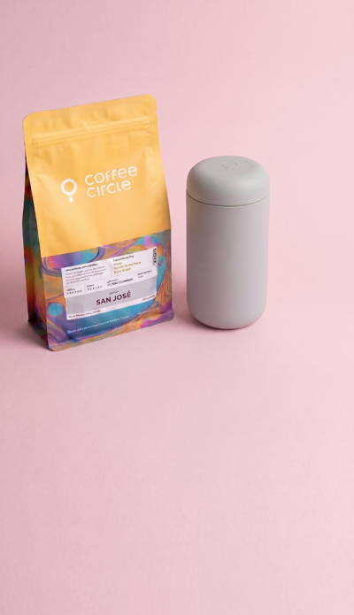A 250 bag from the Coffee Circle brand (variety “San José”) is shown on the left against a pink background. The bag has a yellow upper section with a white logo and a colorful, patterned lower section with a detailed label featuring “SAN JOSÉ” and various scales. To the right of it stands a cylindrical, matte-gray insulated tumbler with a smooth surface.