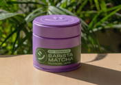 Eine violette Dose mit der Aufschrift „BIO CEREMONIAL BARISTA MATCHA KAGOSHIMA, JAPAN“ steht auf einem hellen Holztisch. Der Hintergrund ist mit üppigen, grünen Pflanzen gefüllt. Das Foto wird von natürlichem Sonnenlicht beleuchtet, das einen Schatten von der Dose auf den Tisch wirft.
