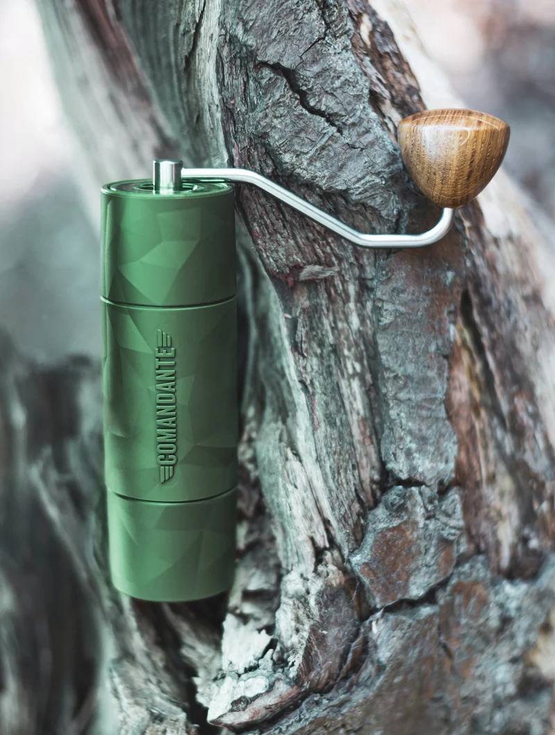 Comandante X25 Trailmaster Forest Kaffeemühle, die an einem groben Baumast lehnt. Die grüne, polygonal-texturierte Mühle mit ihrem Kurbelarm aus Metall und Holzknauf wird von der Rinde eines alten Astes eingerahmt.