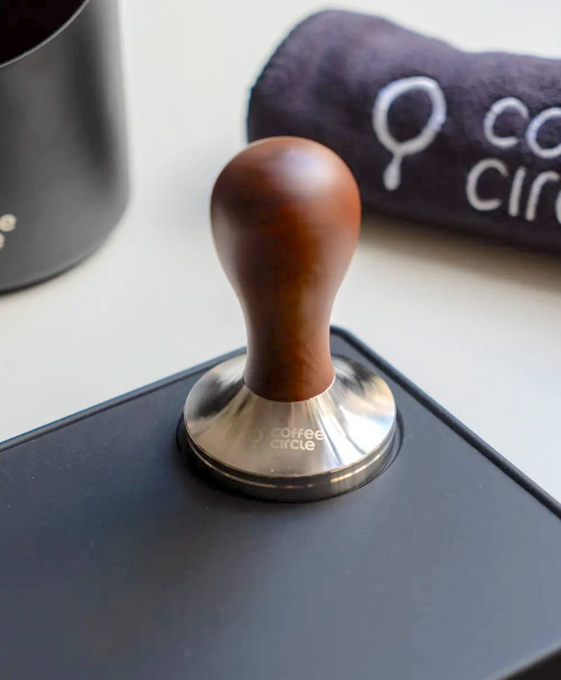 Coffee Circle Tamper Rosenholz ist auf einer Silikonmatte zu sehen. Angrenzend liegt ein Reinigungstuch, welches mit dem Coffee Circle Logo bedruckt ist.