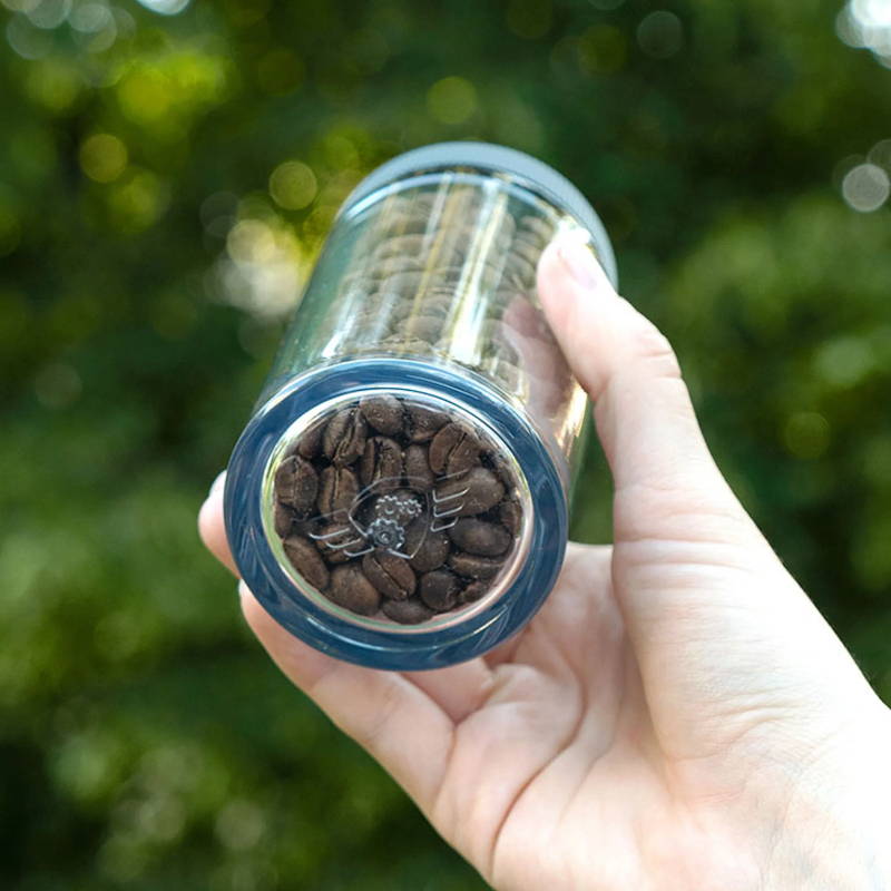 Comandante Polymer large bean container is held in the hand. Behind it, you can see a background of leaves.